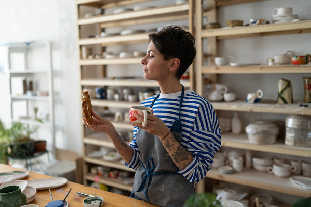 Französische Keramikerin mit Schürze, die ihre Erzeugnisse aus Ton hält, malt und bastelt gern. Junge Frau als Kleinunternehmerin aus Keramik trinkt Tee aus einer handgemachten Tasse. Schöne eigene Arbeit, Hobby-Konzept - Foto, Bild