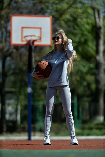 athletic blonde girl stands on the basketball court in a gray tracksuit with a ball in her hands - Foto, immagini