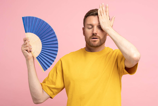 Sweaty tired middle aged man touching forehead using paper fan suffer from heat, feels sluggish. Displeased guy cooling in hot summer weather, isolated on studio pink background. Overheating concept - Photo, Image