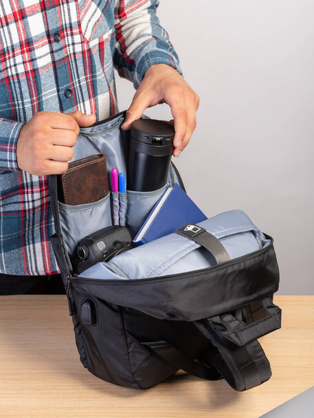 Gros plan d'un jeune étudiant qui sort une tasse à café d'un sac à dos qui contient également un ordinateur portable, un portefeuille, un bloc-notes - Photo, image