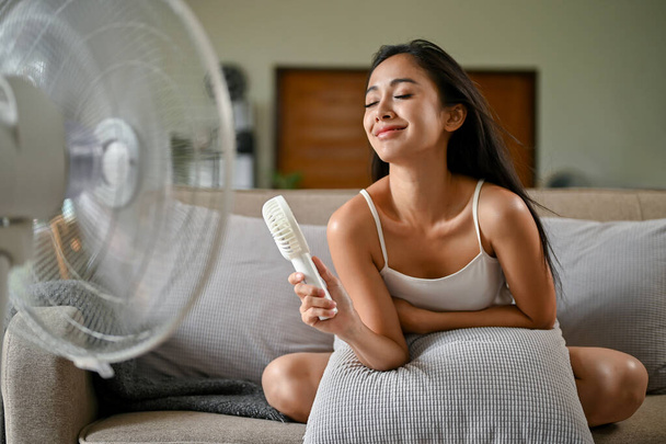 Une femme asiatique magnifique, se sentant chaude souffrant d'une crise de chaleur, se rafraîchit avec un ventilateur pratique et un ventilateur électrique alors qu'elle est assise sur un canapé dans son salon un jour d'été. - Photo, image