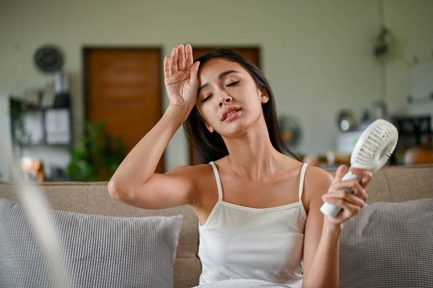 Väsynyt ja ylikuumentunut aasialainen nainen, joka kärsii lämpöhyökkäyksestä, käyttää sähköistä kätevää tuuletinta jäähtymään lepäämällä olohuoneessaan sohvalla kuumana kesäpäivänä.. - Valokuva, kuva