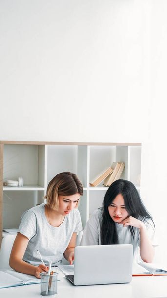 Studying together. Homework cooperation. Two young girls at desk with laptop learning completing task preparing for exam writing notes at home empty space. - 写真・画像