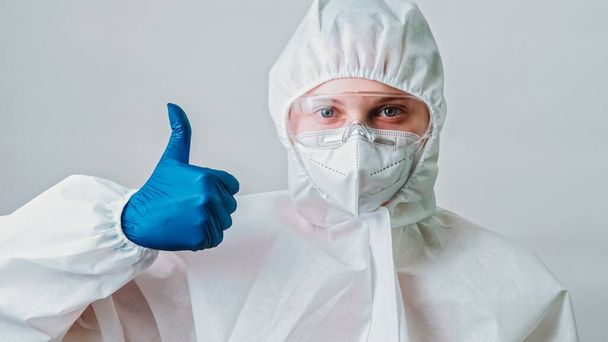 Medical virologist. Covid-19 protective equipment. Approval gesture. Female lab specialist nurse woman in white coverall suit thumb up hand sign isolated on grey background. - 写真・画像