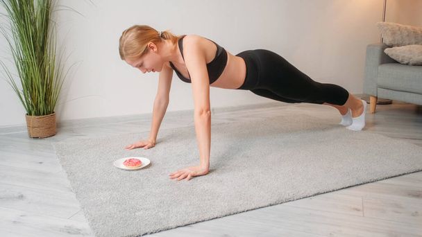 Fitness motivation. Weight loss diet. Calorie burn. Confident athletic woman performing push-up exercise on floor with sweet doughnut dessert. - Foto, immagini