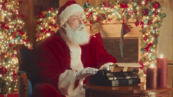 Medium slowmo of pensive kind Santa in red hat and robe typing letter to children on typewriter, sitting in his chair in beautifully decorated room with sparkling lights, Christmas tree and fireplace - Felvétel, videó