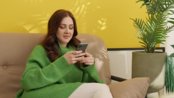 Smiling pretty brunette browsing cell phone, sitting on sofa with crossed legs. Young woman types on cell phone, biting her lip seductively, flirting. High quality 4k footage - Footage, Video