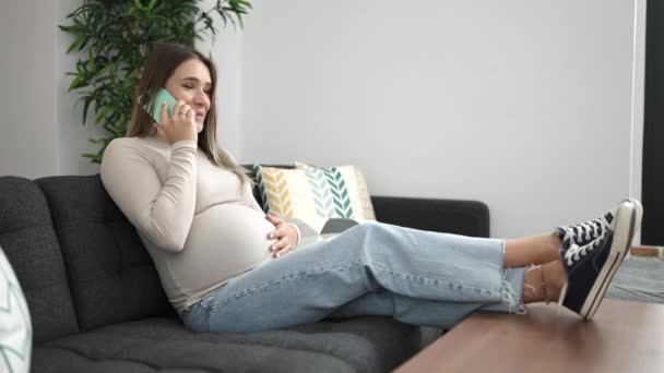 Νεαρή έγκυος γυναίκα που μιλάει στο smartphone αγγίζοντας την κοιλιά στο σπίτι - Πλάνα, βίντεο