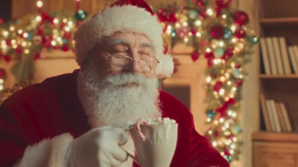 Medium closeup with slowmo of happy Santa Claus in red costume drinking hot chocolate with marshmallows from mug, sitting in his chair by fireplace decorated with shining lights - Imágenes, Vídeo