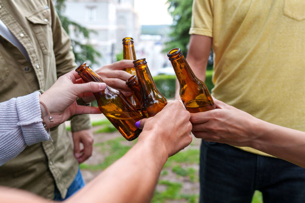 Cinco amigos anónimos en un parque de la ciudad, sus manos tintineando vasos de cerveza en un gesto simbólico de camaradería y celebración. - Foto, Imagen