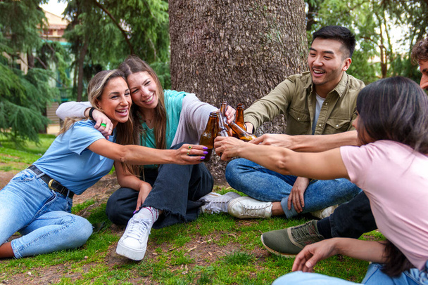 5人の多文化の友人たちが喜びの瞬間を共有し、街の公園に座って、お祝いと幸福のビールグラスを上げます。. - 写真・画像