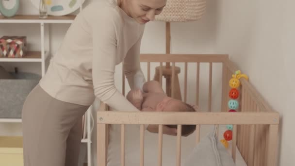 Medium slowmo of young Caucasian woman putting her beautiful newborn baby daughter in wooden crib to sleep - Filmati, video