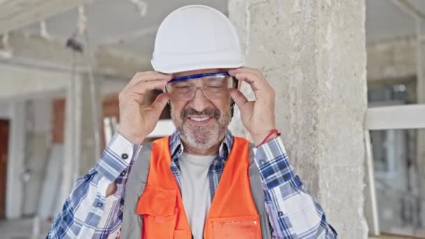 Будівельник середнього віку посміхається впевнено виймаючи окуляри на будівельному майданчику - Кадри, відео