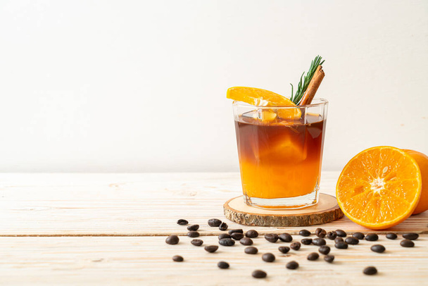 Sklenice ledové americké černé kávy a vrstva pomerančové a citrónové šťávy zdobené rozmarýnem a skořicí na dřevěném pozadí - Fotografie, Obrázek