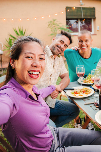 Vertical portrait of a group of mature adult best friends smiling taking a selfie on a barbeque party celebration at home backyard. Middle age happy people shooting pictures sitting at patio table - Zdjęcie, obraz