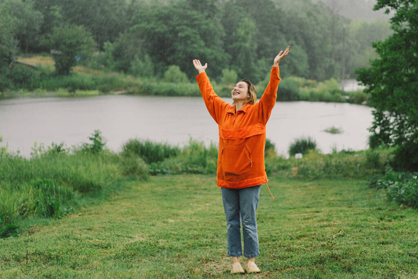 Πορτρέτο ενός νεαρού κοριτσιού με πορτοκαλί πουλόβερ που απολαμβάνει τη βροχή μια καλοκαιρινή μέρα. Το κορίτσι διασκεδάζει και χορεύει κάτω από τη καλοκαιρινή βροχή. - Φωτογραφία, εικόνα