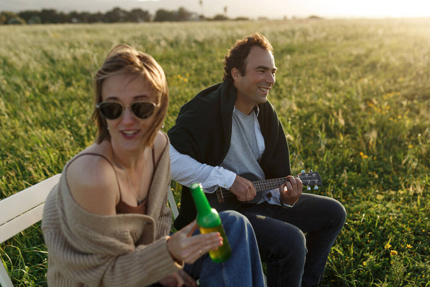 Summer time. Beautiful young couple enjoying time in field around grass. Lifestyle, love, dating, vacation concept. Private. Woman drinking beer, man playing ukulele  - Фото, зображення