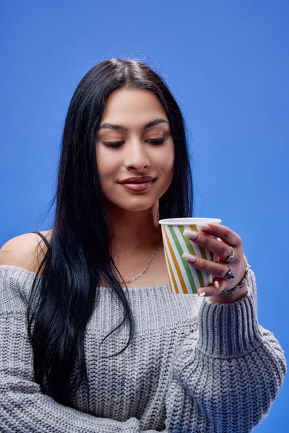 Портрет красивой юной брюнетки, позирующей в студии на синем фоне с чашкой кофе в руке - Фото, изображение