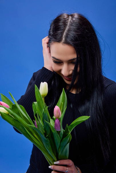 Eine schöne junge Frau mit Tulpen in der Hand posiert im Studio vor blauem Hintergrund - Foto, Bild