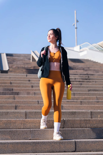 vertical photo of a beautiful caucasian sportswoman smiling walking down urban stairs, concept of active and sportive lifestyle, copy space for text - Foto, imagen