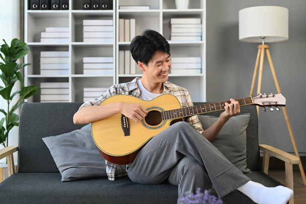 Fröhlicher junger asiatischer Mann spielt Akustikgitarre auf Couch und verbringt Freizeit im gemütlichen Wohnzimmer. - Foto, Bild