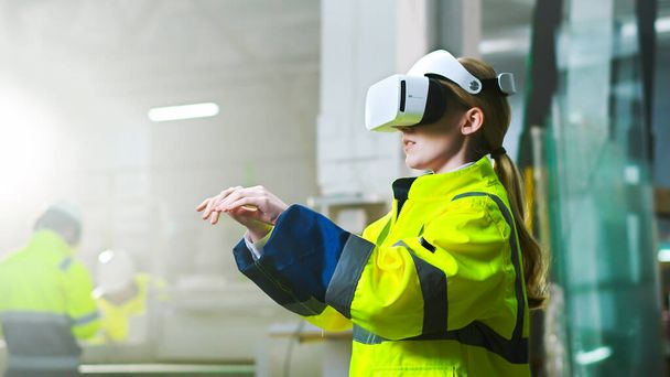 Caucasian female young engineer in headset and yellow uniform standing in factory and having virtual reality experience. Engineering concept. Inside. Blond woman in VR goggles moving hands at plant. - Foto, afbeelding