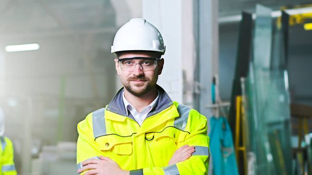Portrait of handsome Caucasian man in helmet and uniform standing at plant manufactoring and taking on goggles. Indoors. Young worker at factory in hardhat and protectional glasses. Protection. - Фото, зображення