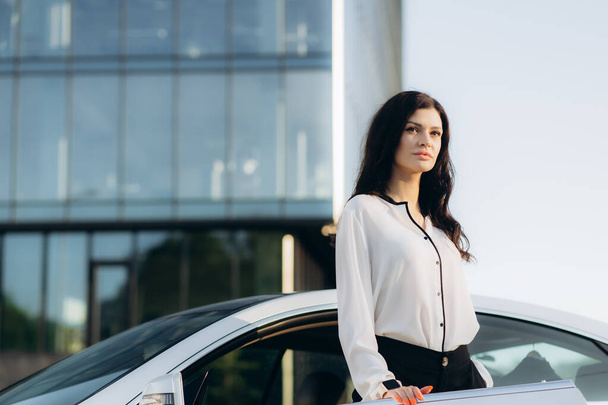 Portrait of confident stylish businesswoman holding mobile phone looking at camera standing near car, copy space. Transportation concept - Photo, image