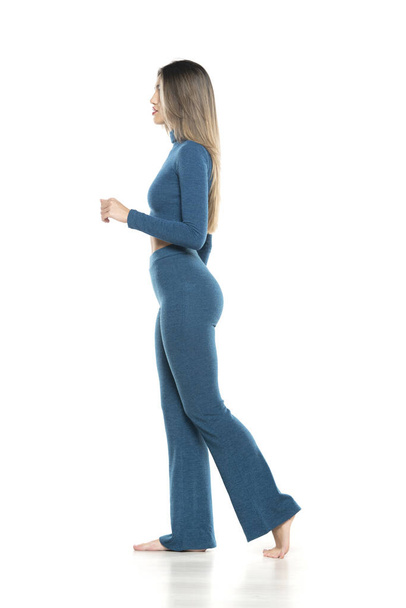 young modern barefeet woman in blue pants and blouse walking on white studio background. side, profile view - Foto, afbeelding