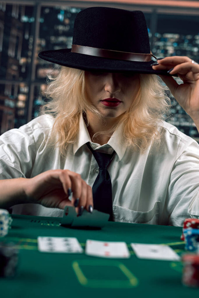 ελκυστική κοπέλα με πουκάμισο και καπέλο που καλύπτει το πρόσωπο με καπέλο κρατώντας κάρτες πόκερ στο καζίνο. πόκερ. Κυρία παίχτρια - Φωτογραφία, εικόνα
