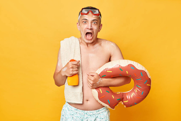 Overjoyed smiling happy tourist man at summer resort standing with rubber ring isolated over yellow background looking at camera with open mouth being amazed. - Fotó, kép