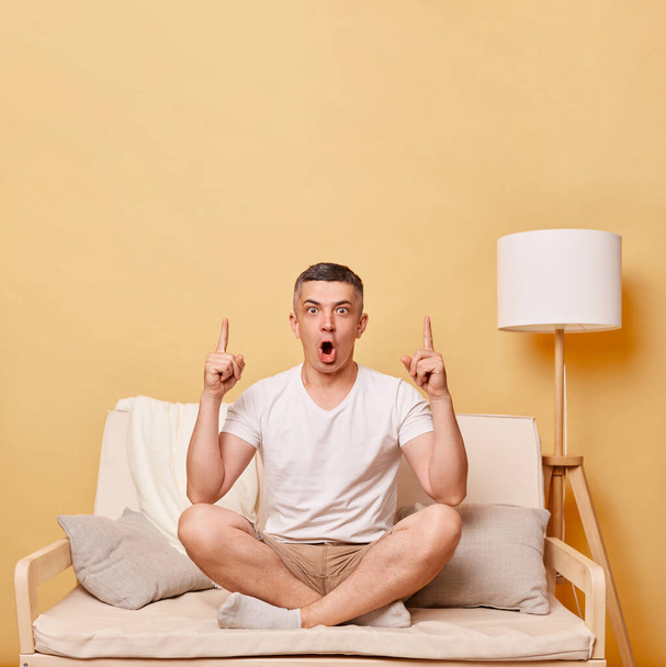 Exited surprised astonished man wearing white T-shirt sitting on sofa isolated over beige background pointing upwards at advertisement area space for promotional text. - Fotó, kép