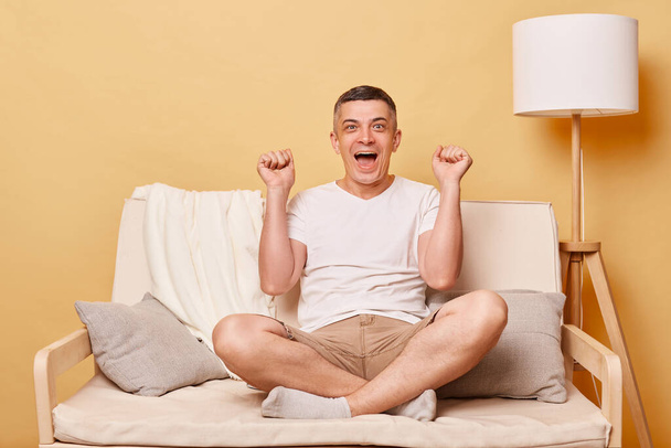 Extremely happy joyful man wearing white T-shirt sitting on sofa isolated over beige background clenched fists with overjoyed expression celebrating his success screaming happily. - Photo, Image