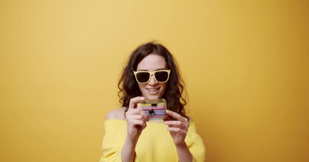 Een jong meisje lacht en maakt een foto met de retro camera op een gele achtergrond, ze is gelukkig. hobby-activiteit. Zelffoto. Stuur bericht on line. Zomer Relax. Fijne reis. Leuke foto. - Video