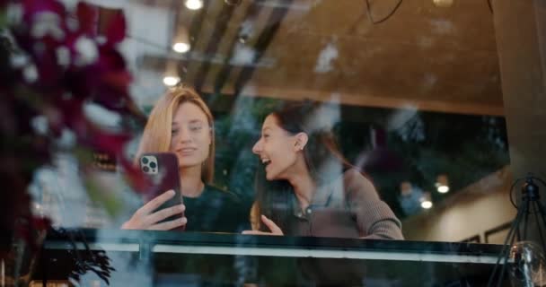 Jonge vrouwen die selfie nemen in het restaurant. Low angle handheld schot van jonge brunette tonen tong terwijl het nemen van selfie met gelukkig vriendin achter glas in restaurant - Video