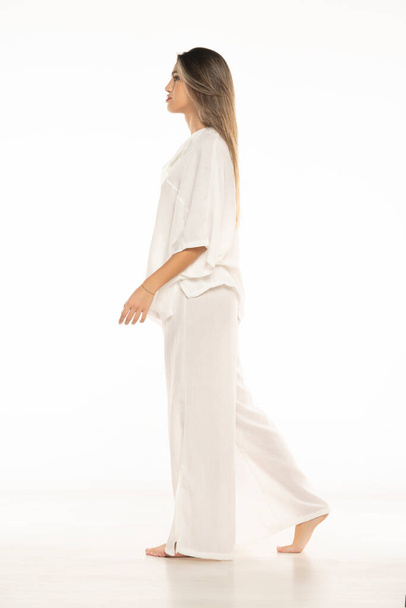 mladá moderní bosá žena v bílých kalhotách a halence chůze na bílém pozadí studia. boční pohled - Fotografie, Obrázek