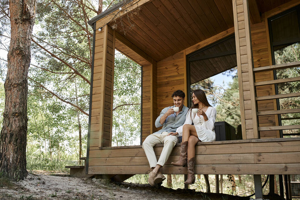 happy couple holding cups of coffee, man and woman smiling on porch of vacation house in forest - Фото, зображення