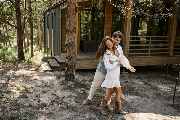 happy man hugging woman and smiling near vacation house, forest, summer and romance, couple in love - Фото, зображення