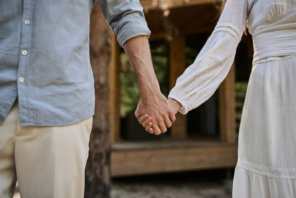przycięty widok, kobieta w sukience trzyma się za ręce z mężczyzną, zamazany letni dom, miłość i romans - Zdjęcie, obraz