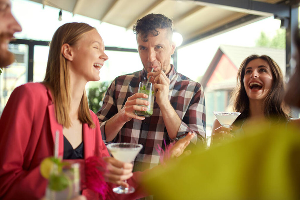 Joyful young people, men and women, friends meeting at cafe, drinking cocktails, talking, having good time together. Concept of friendship, meeting, fun, communication, alcohol drinks - Φωτογραφία, εικόνα