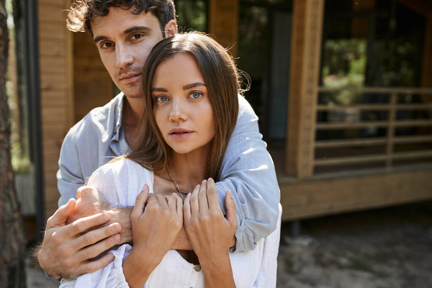 Brunette man hugging girlfriend and looking at camera near blurred vacation house outdoors - Photo, Image