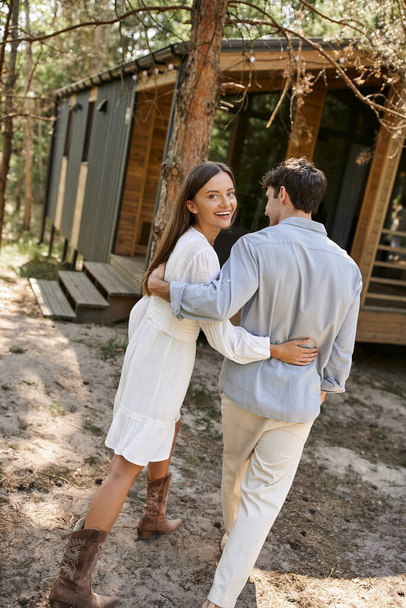 Trendy and smiling woman in sundress hugging boyfriend and looking at camera near vacation house - Foto, Bild