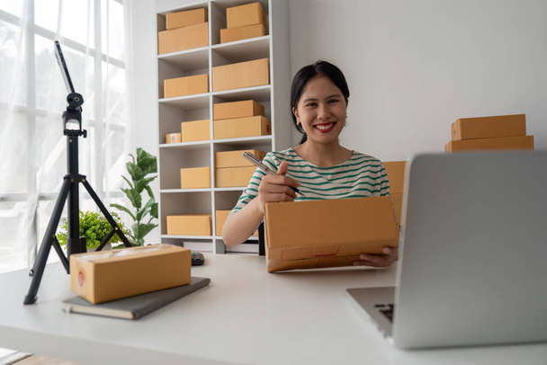 Young business woman working online e-commerce shopping at her shop. Young woman seller prepare parcel box of product for deliver to customer. Online selling, e-commerce. - 写真・画像