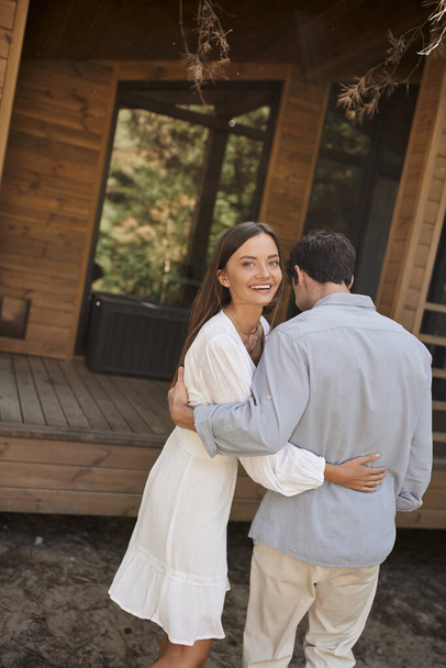 Joyful young woman in sundress hugging boyfriend and looking at camera near vacation house outdoors - Photo, Image