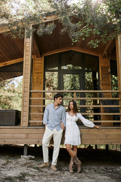 Trendy romantic couple in casual clothes standing under trees near vacation house outdoors - Photo, Image