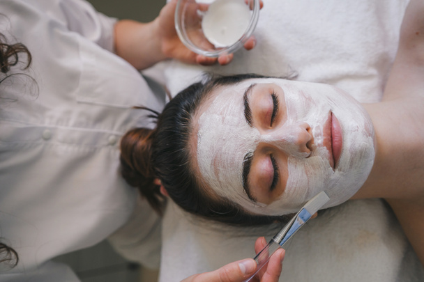 Blanke vrouw doet schoonheidsbehandeling in spa salon en wordt crème op haar gezicht aangebracht. Gezicht peeling masker, spa schoonheidsbehandeling, huidverzorging. - Foto, afbeelding