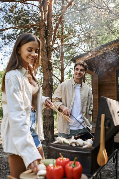 Smiling man cooking on barbecue near blurred girlfriend with vegetables and vacation house outdoors - Zdjęcie, obraz