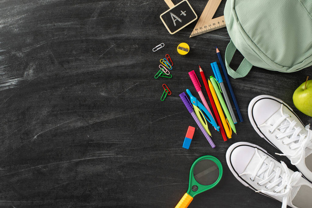 Learning essentials arrangement. Overhead view image featuring a backpack, footwear and an abundance of school supplies on an isolated blackboard backdrop, perfect for text or advertising placement - Photo, Image