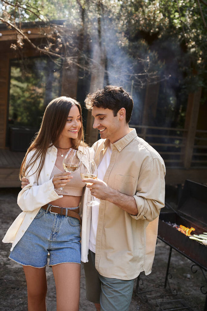 Couple souriant tenant du vin près du barbecue flou et de la maison de vacances en arrière-plan à l'extérieur - Photo, image