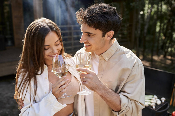 Allegro uomo abbracciare la ragazza con vino vicino bbq e casa di vacanza sullo sfondo - Foto, immagini
